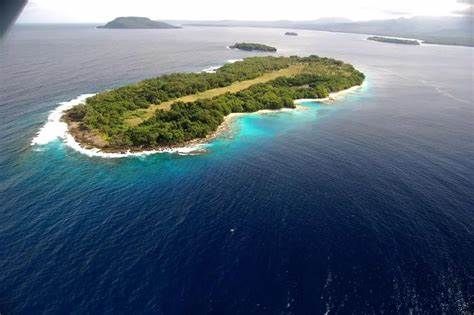 Aerial view of a small lush green island surrounded by deep blue ocean and turquoise shallows.