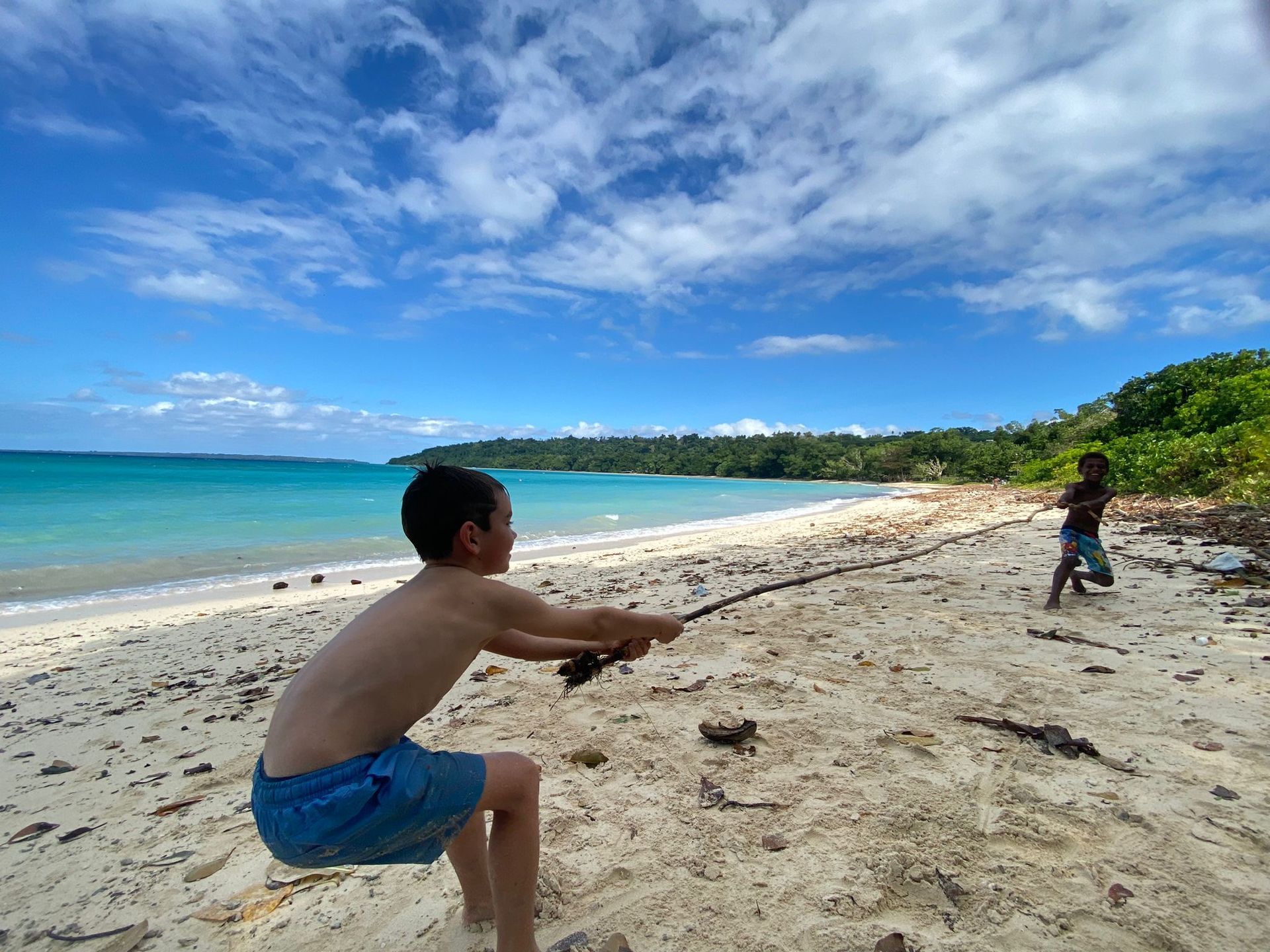 Two people on a sandy beach by turquoise water, one crouching and tossing something toward the other.