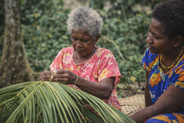 Two people weaving long green palm fronds outdoors in a rural setting