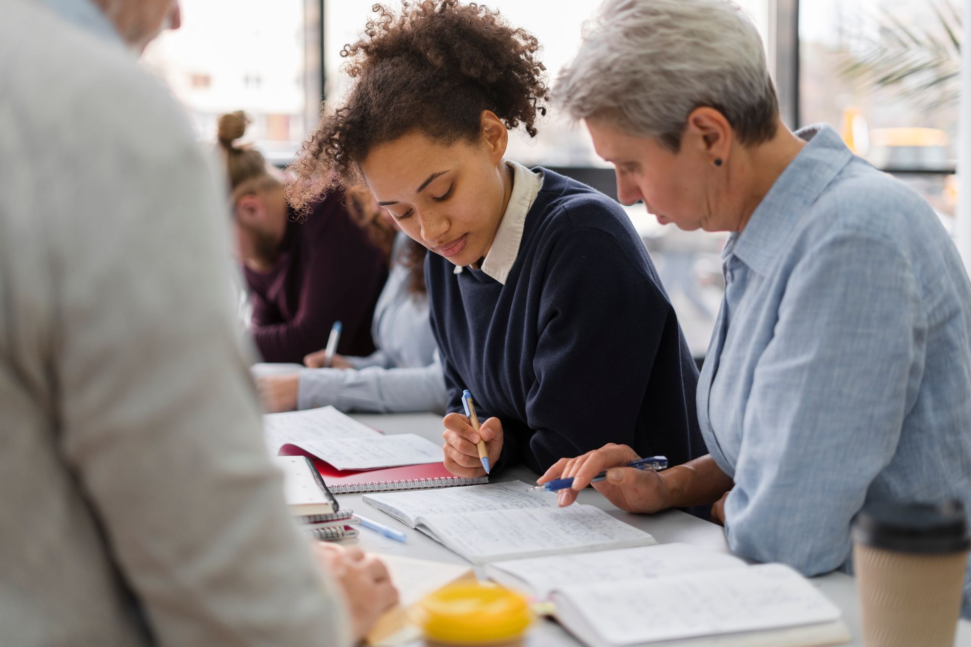 Woman with curly hair and older woman study together, writing in notebooks at a table.