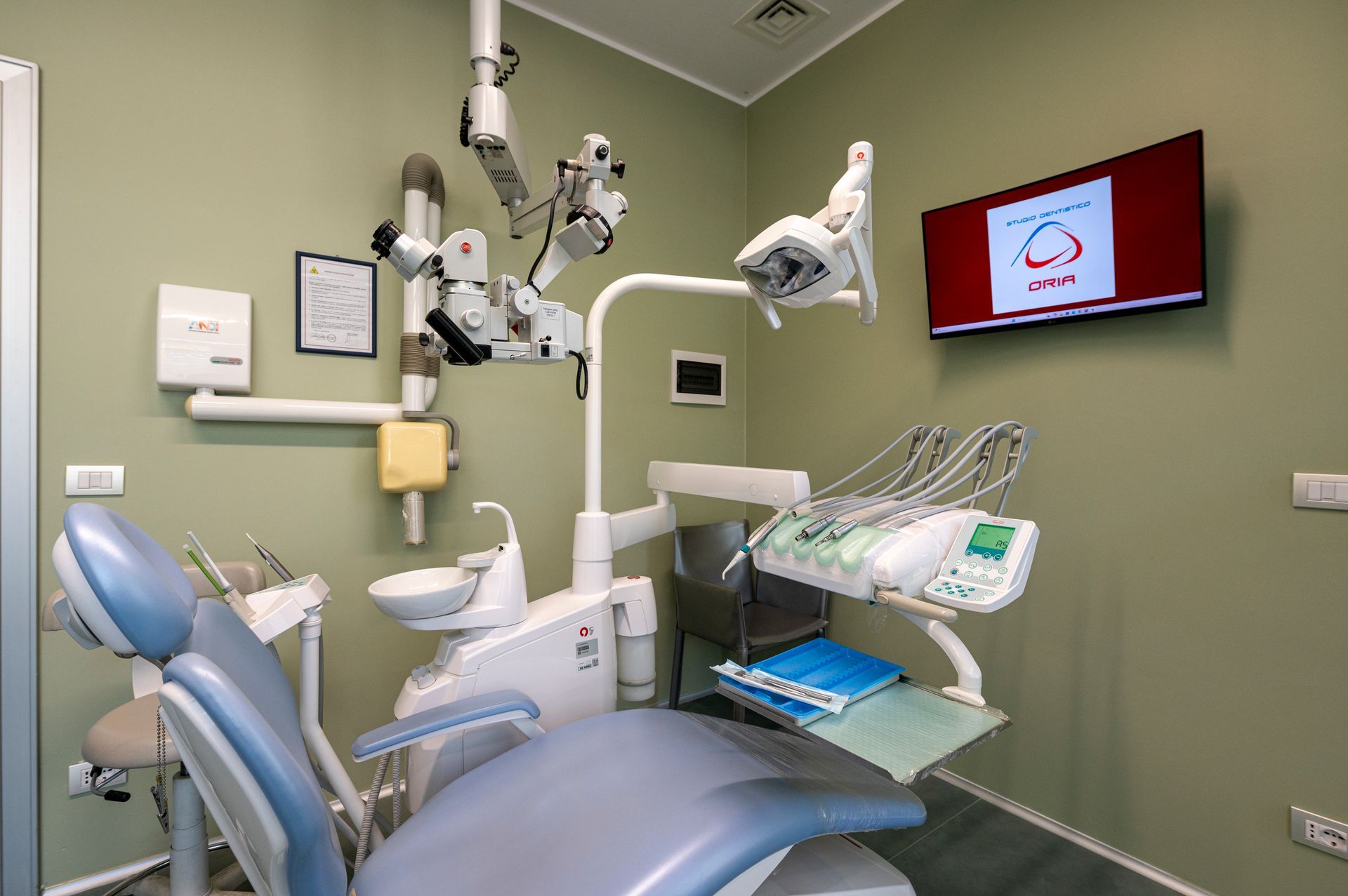 Studio dentistico con poltrona, luci e attrezzatura. Una TV con logo e pareti dai toni neutri.