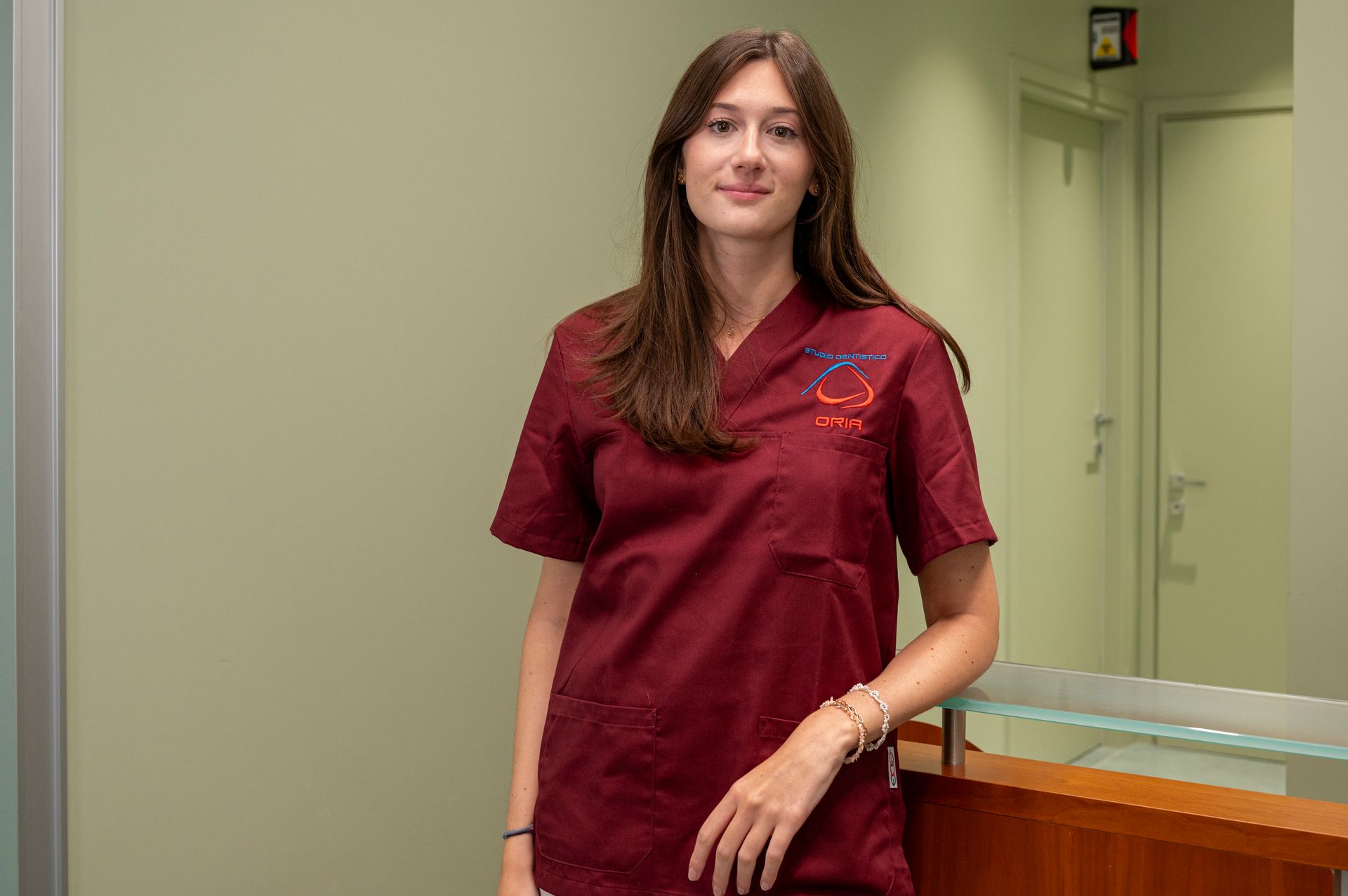 Una donna in camice color castagna è in piedi vicino al bancone di uno studio medico e sorride leggermente.