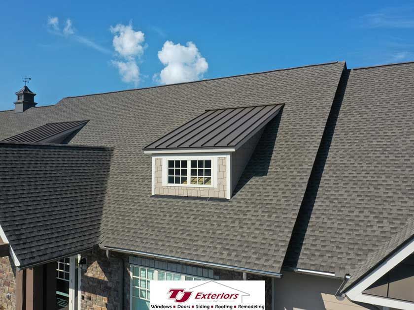 Gray shingle roof with a dormer and blue sky. TJ Exteriors logo at the bottom.