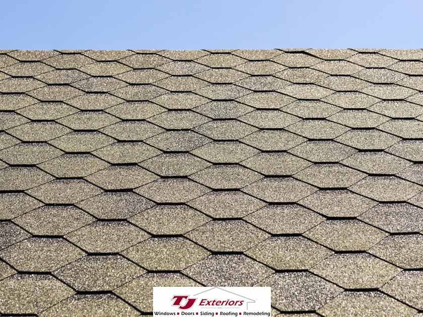 Close-up of a roof covered in hexagonal asphalt shingles in shades of brown and tan against a blue sky.