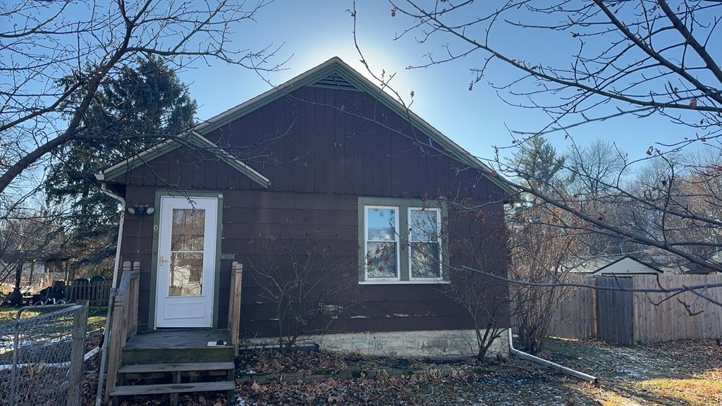 A small brown house with a white door and windows is surrounded by trees.
