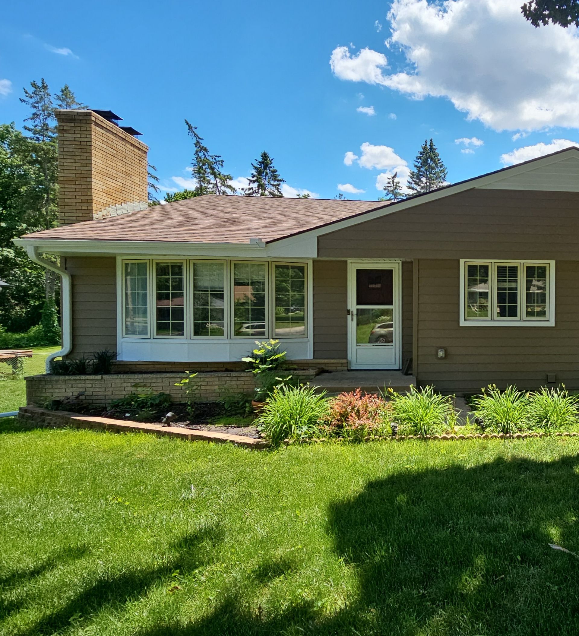 Single-story suburban house with tan siding, white-trim windows, and a green lawn under a blue sky