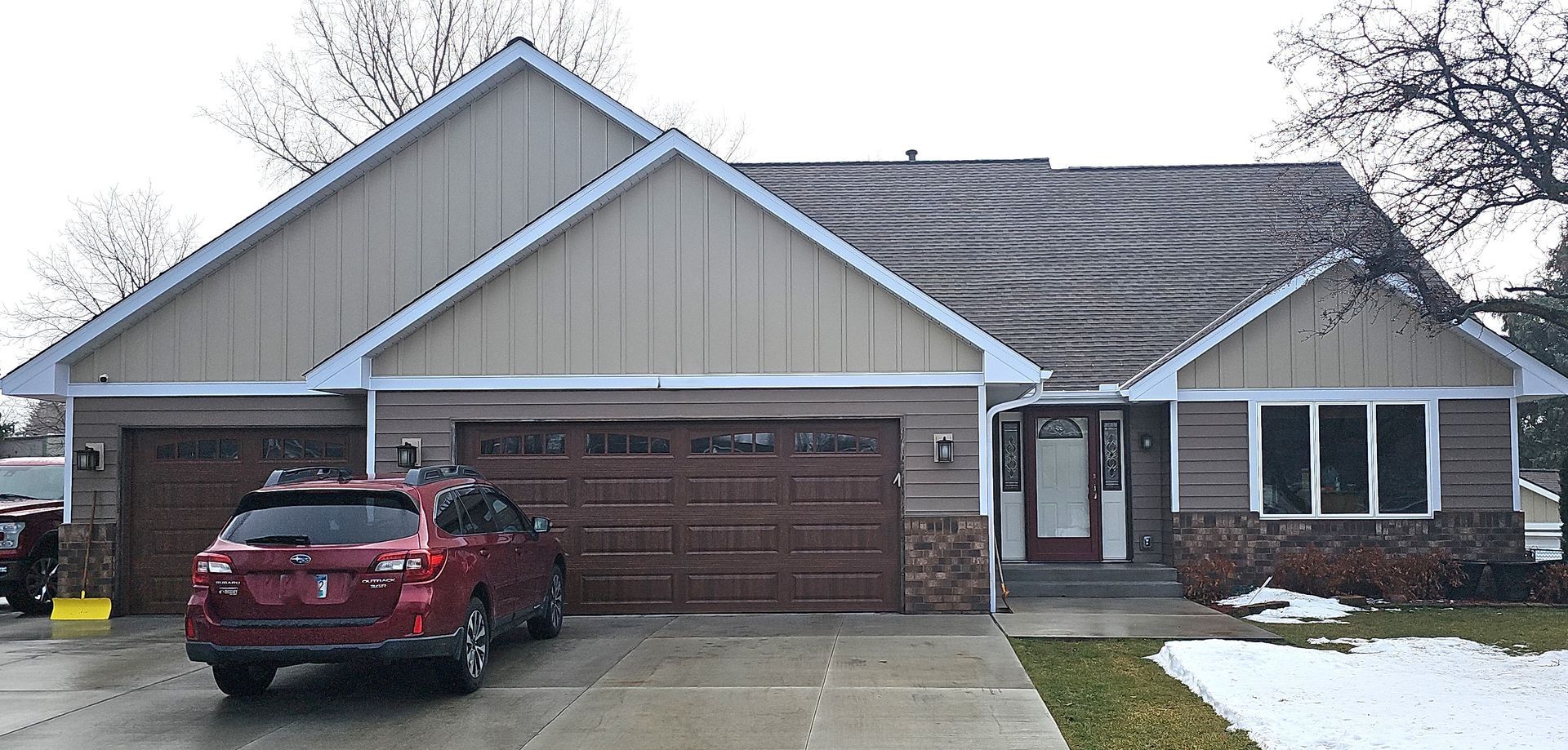 Snow-covered suburban house with a red SUV parked in the driveway and a double garage.