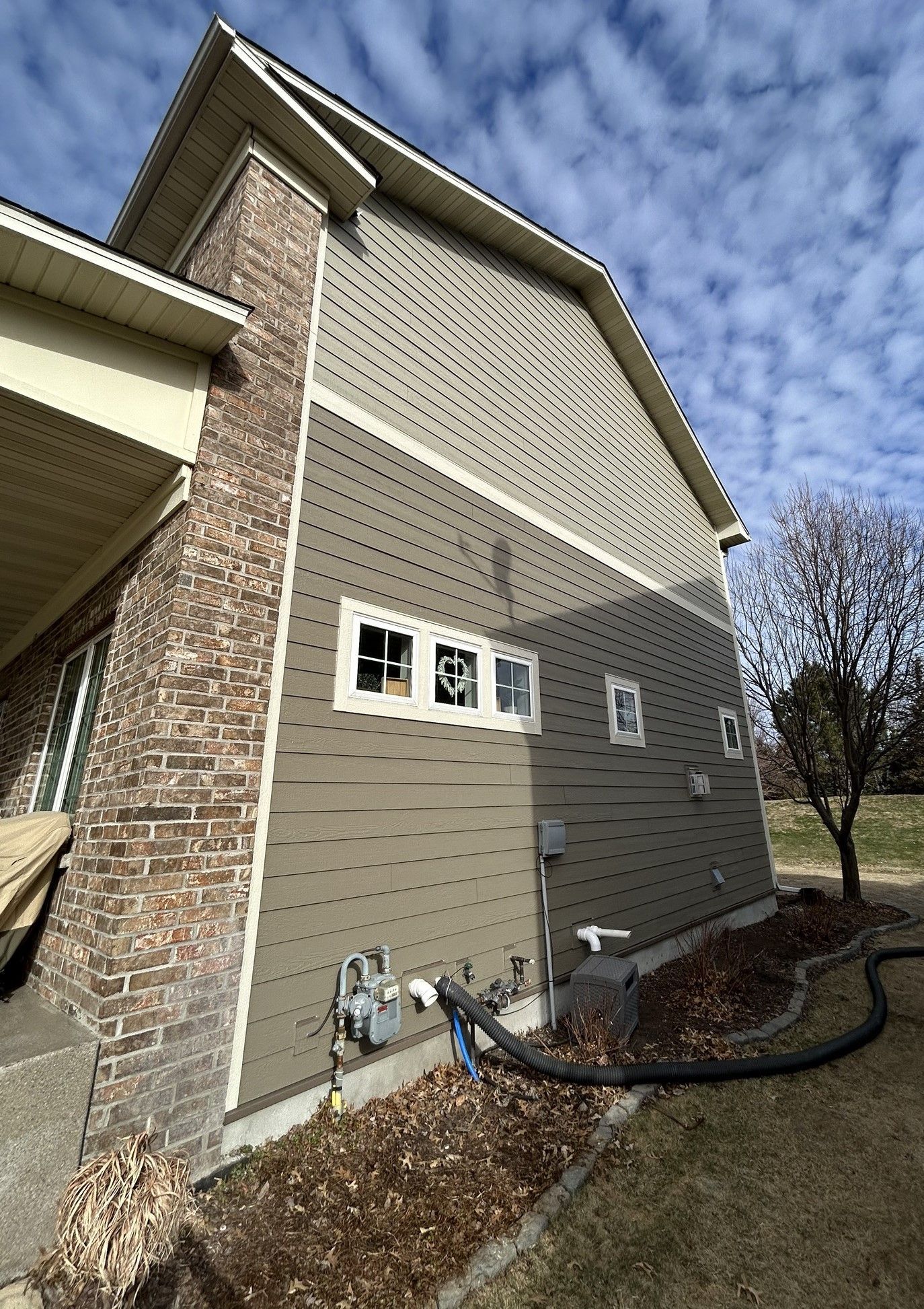Exterior of a brick and siding house with utility meters and hoses along the foundation under a cloudy sky
