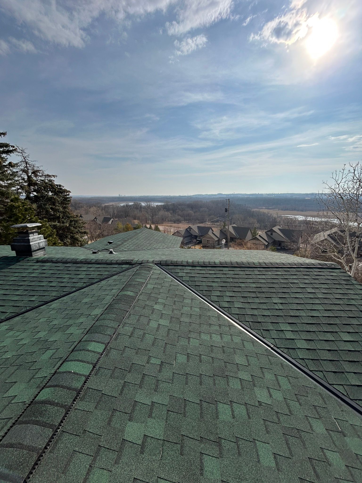View from a green-shingled rooftop over trees and a distant city skyline under a bright blue sky
