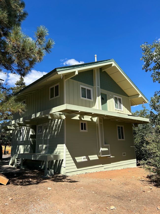 Two-story beige house with green trim under a clear blue sky, surrounded by trees and dry ground