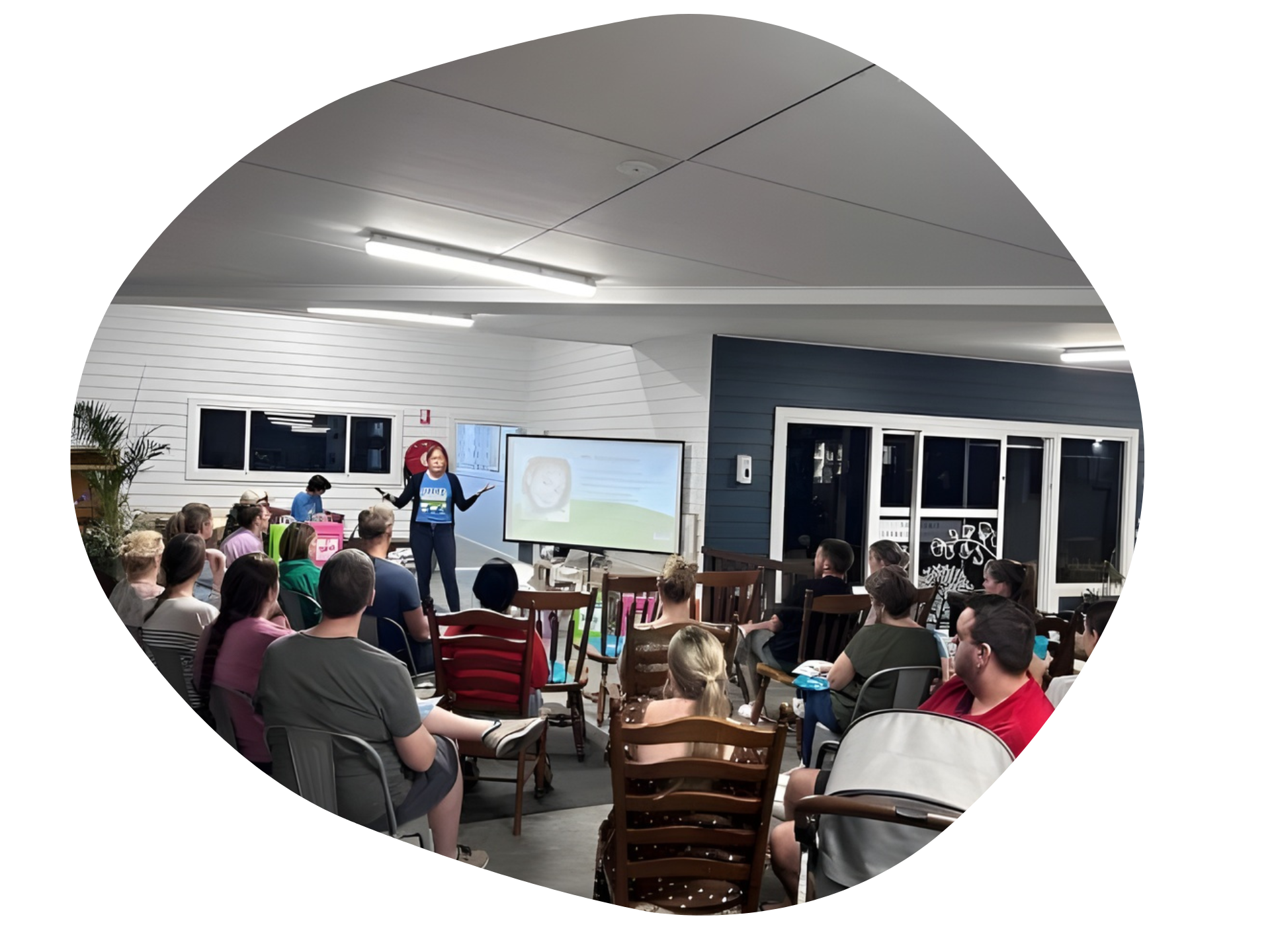 Person presenting to a seated audience in a room; a screen and windows are visible.