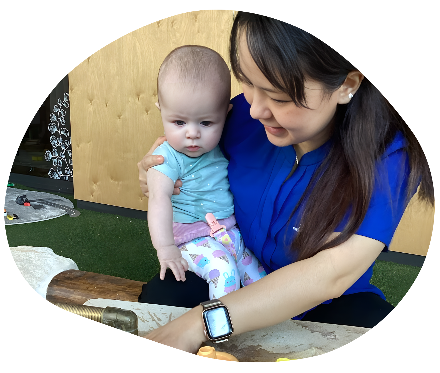 Woman holding baby outside. They sit at a table. Baby wears colorful pants.