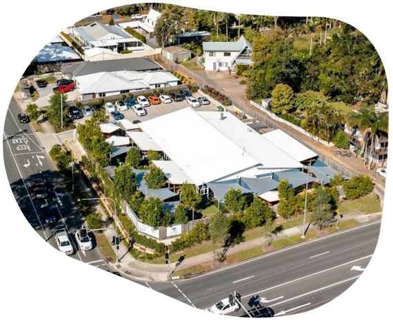 Aerial view of a white-roofed building surrounded by trees and a parking lot, near a road with cars.