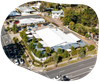 Aerial view of a white-roofed building and surrounding structures, with a parking lot and a road lined with cars and trees.