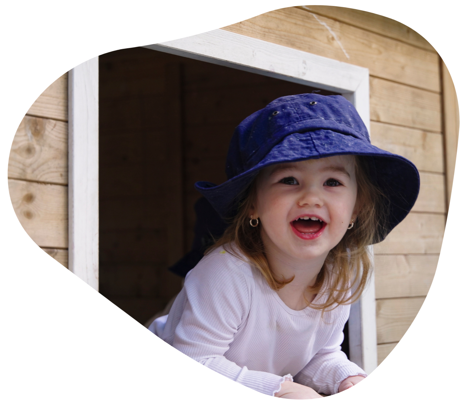 Young child in blue hat smiles from wooden window.