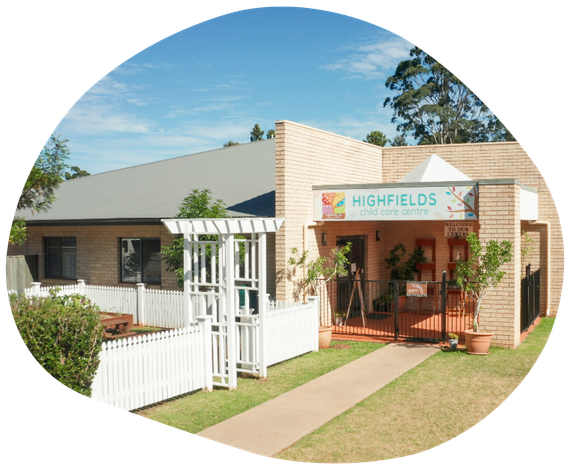 Highfields Early Learning Centre exterior: tan brick building with sign, entrance, picket fence, and greenery.
