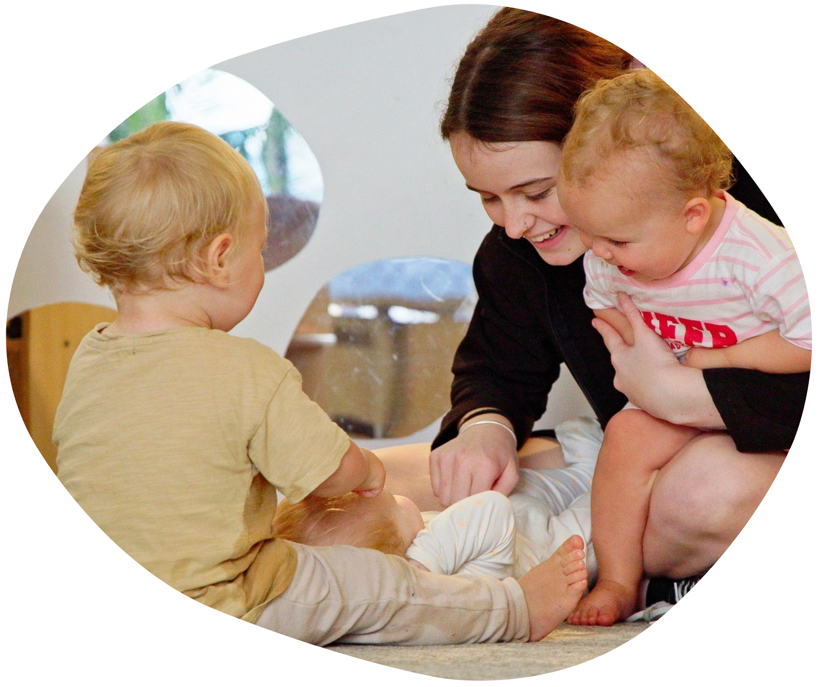 A caregiver changes a baby's diaper as two toddlers look on, all smiling and in a light-filled room.