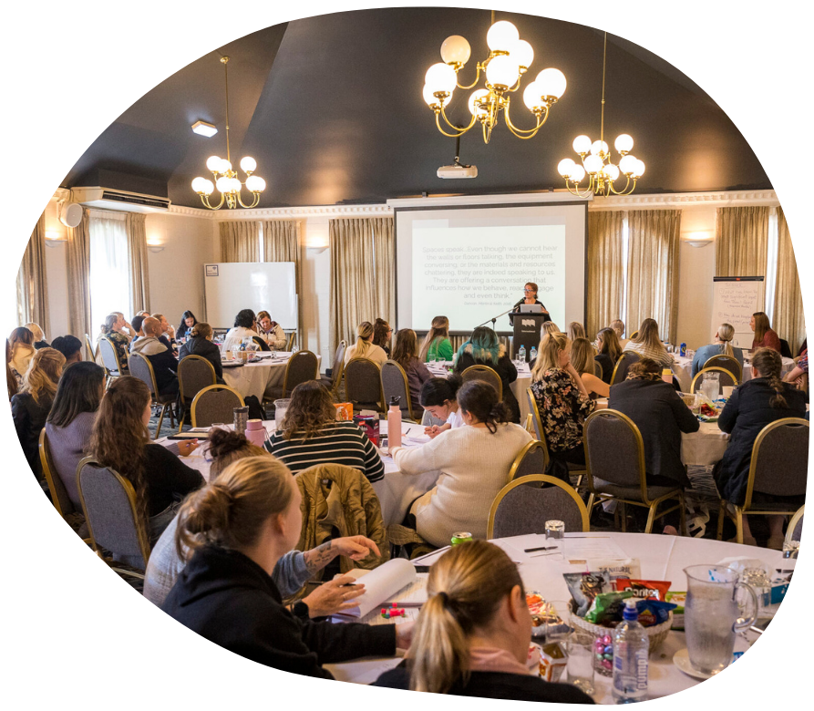 Conference room with presenter, audience seated at round tables, and a projection screen.
