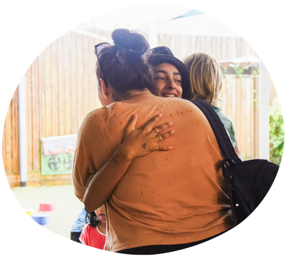 People hugging, smiling in front of a wooden fence; someone has a bag.