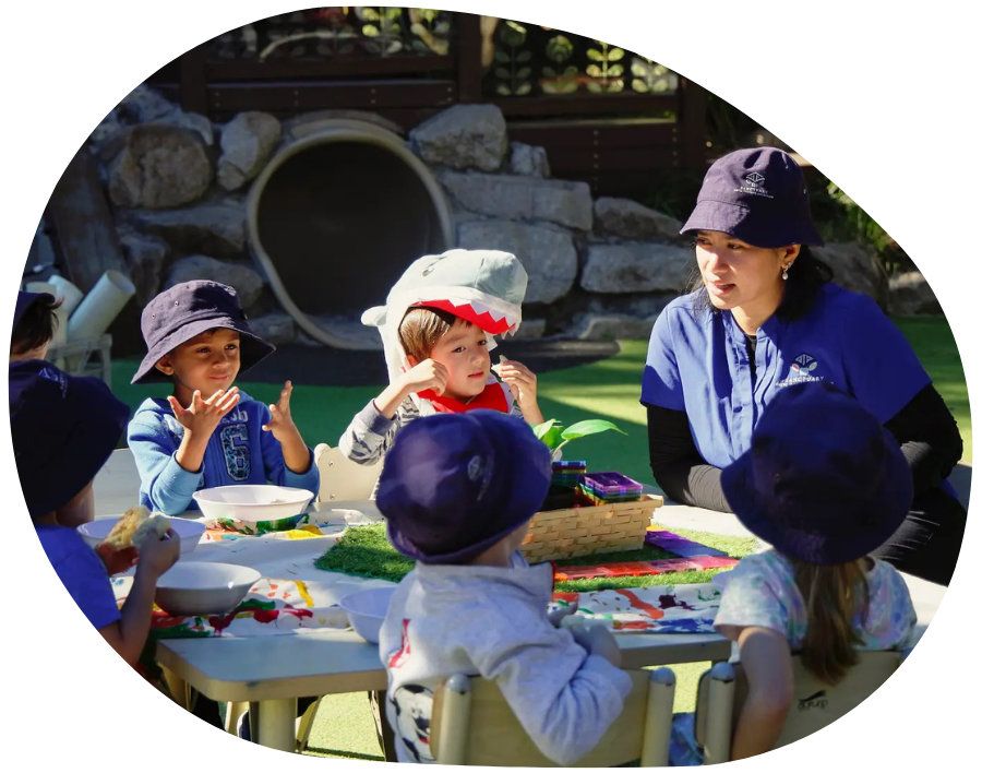 Children and teacher seated around a table, wearing blue hats. One child in a shark hat. Outdoor setting.