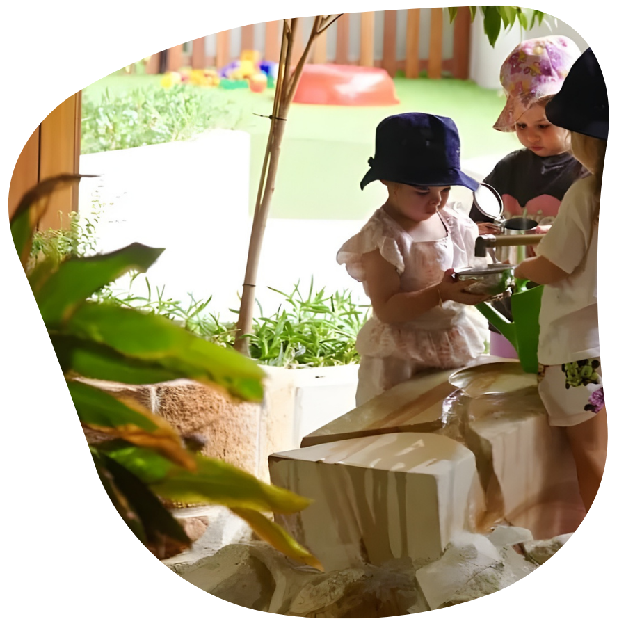 Children playing with water in an outdoor setting; one holds a bowl, others watch, greenery in foreground.