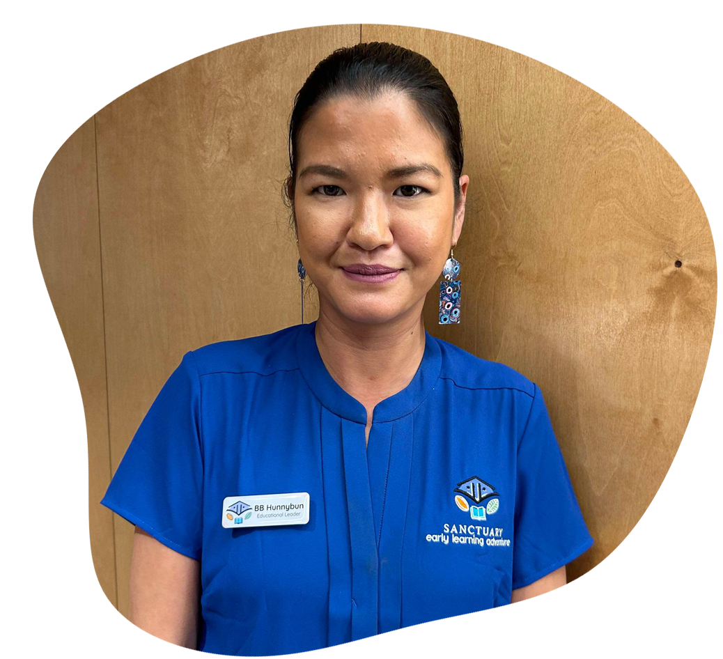 Woman in blue scrubs smiles, name tag visible. Wooden panel background.