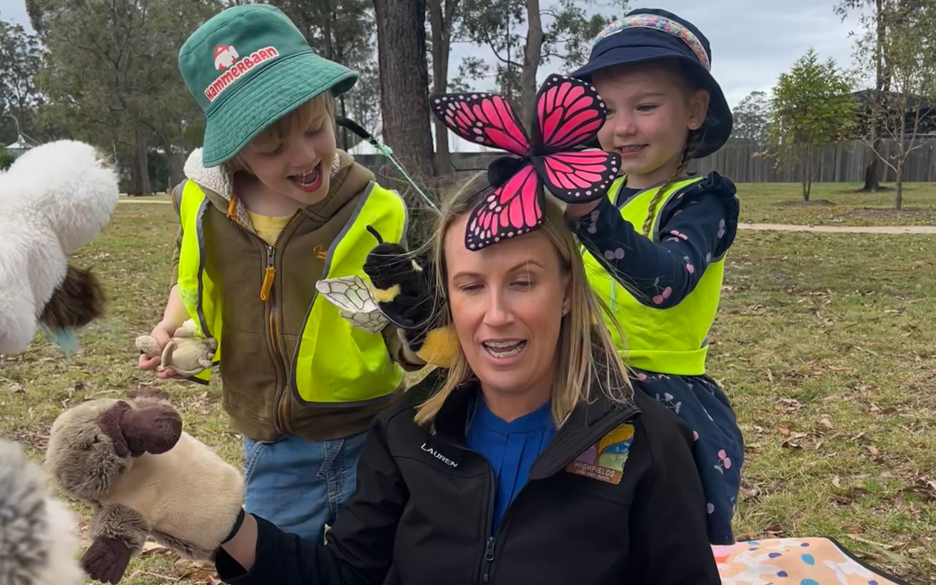 Two children in high vis vests stand behind a woman who is sitting on the ground. 