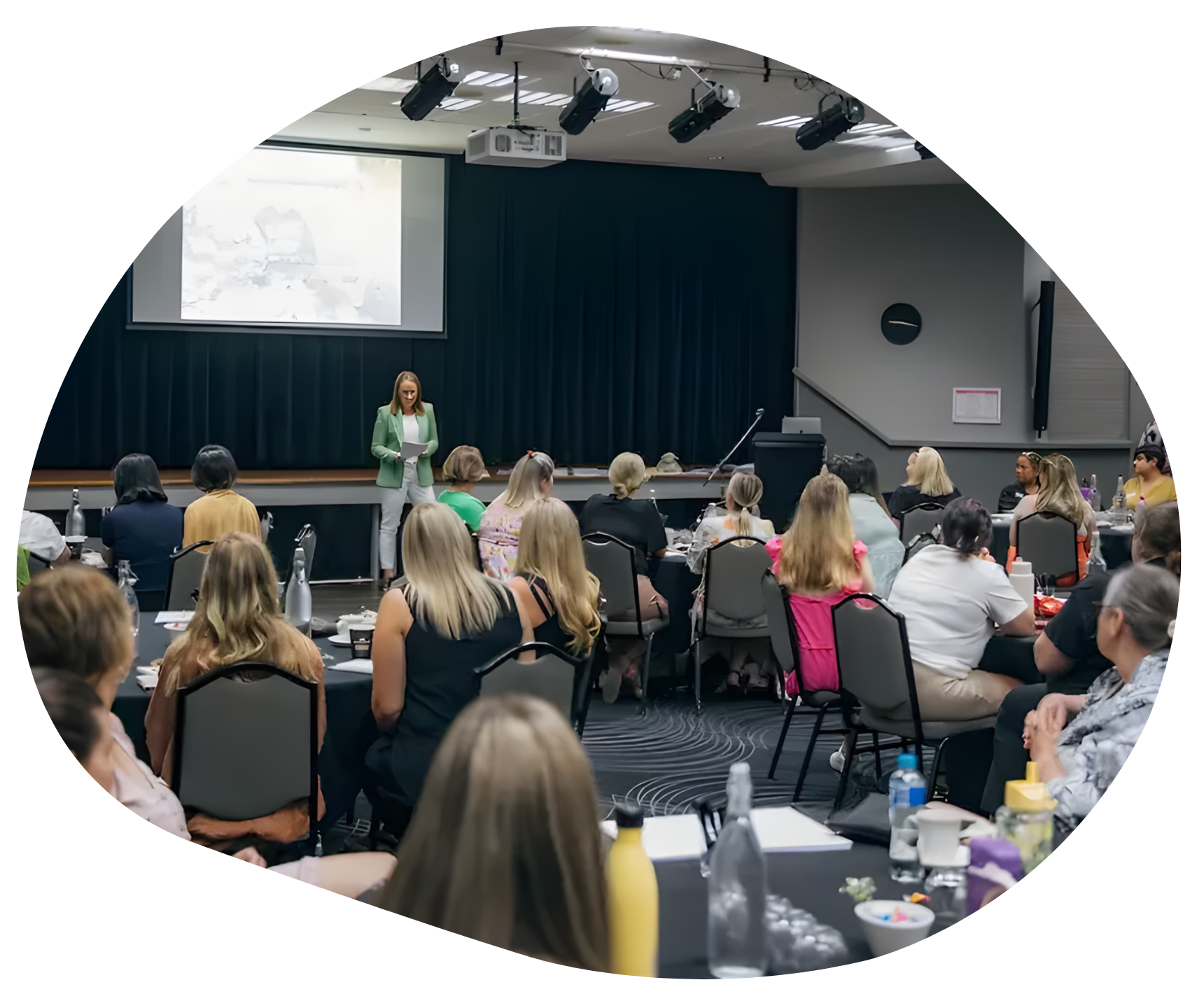 Woman presenting to audience in a conference room. Attendees seated at tables, screen with presentation.