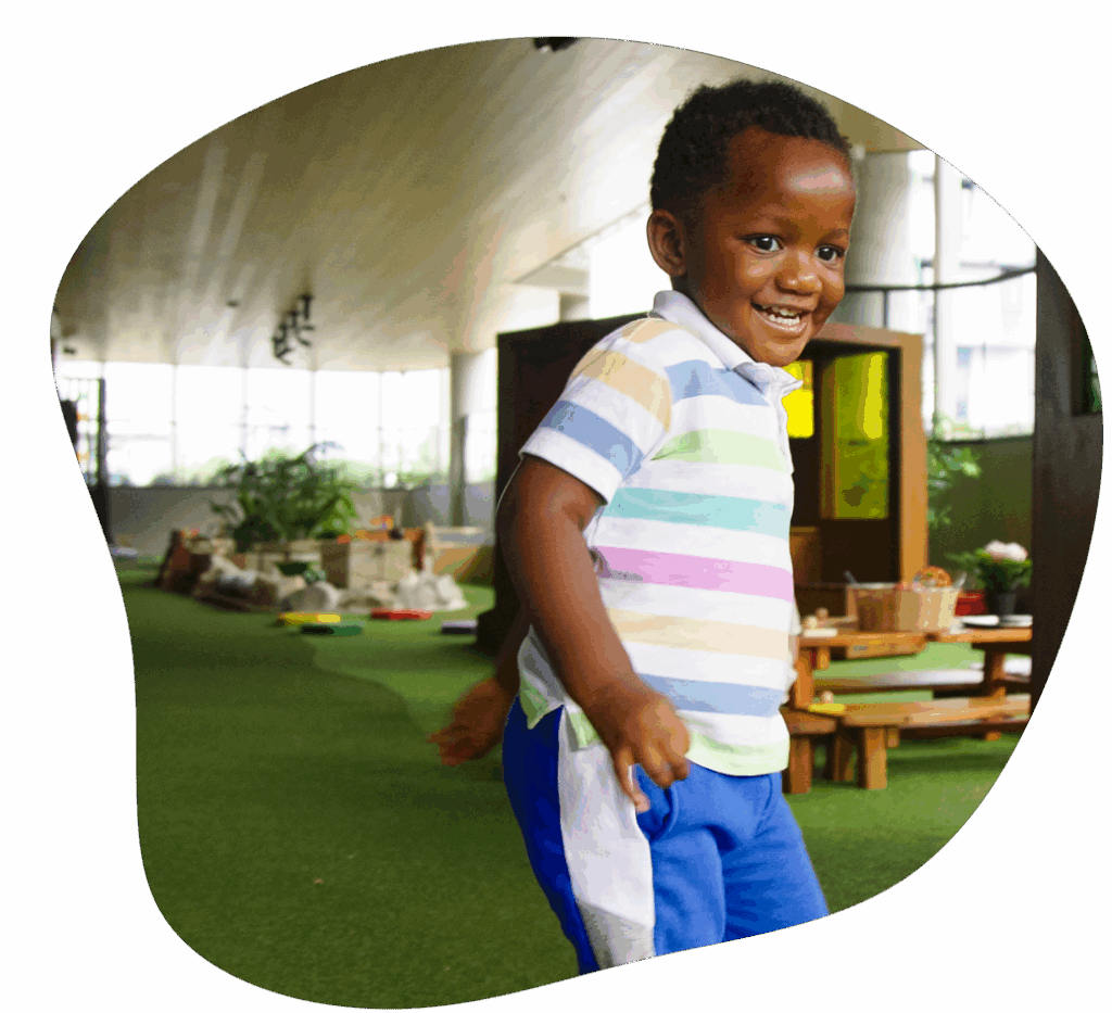 Boy in striped shirt and blue pants smiles, running indoors on green carpet.
