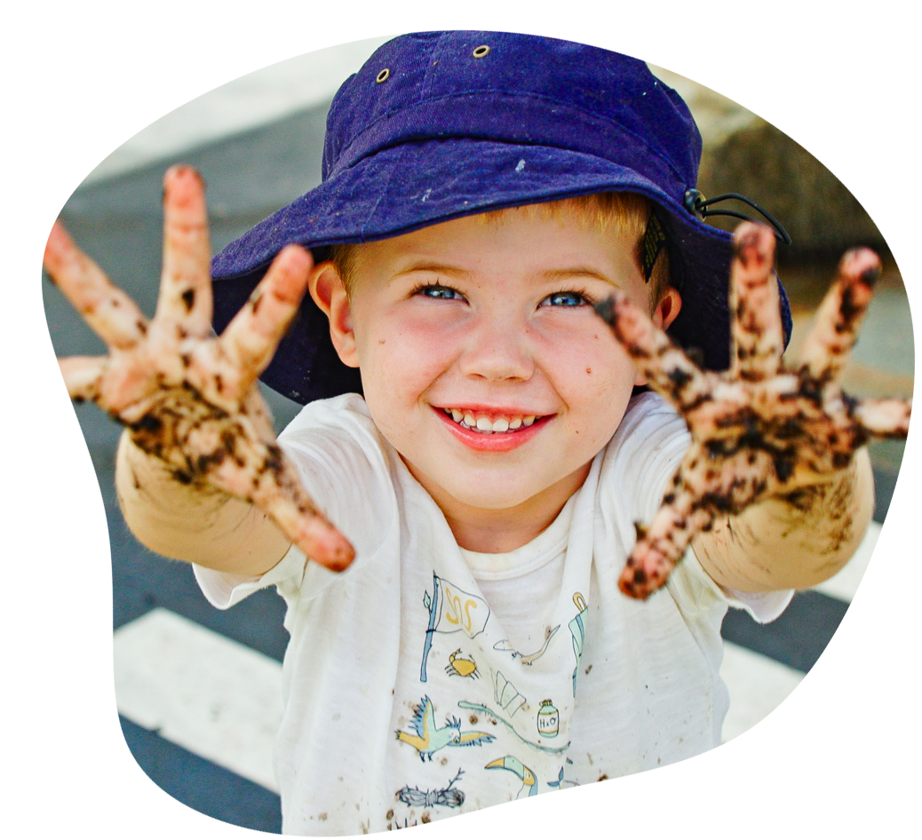 Boy with a blue hat and muddy hands smiles, arms outstretched.