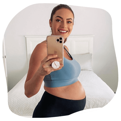 Pregnant person takes selfie in bedroom, wearing blue sports bra, black pants, and smiling.
