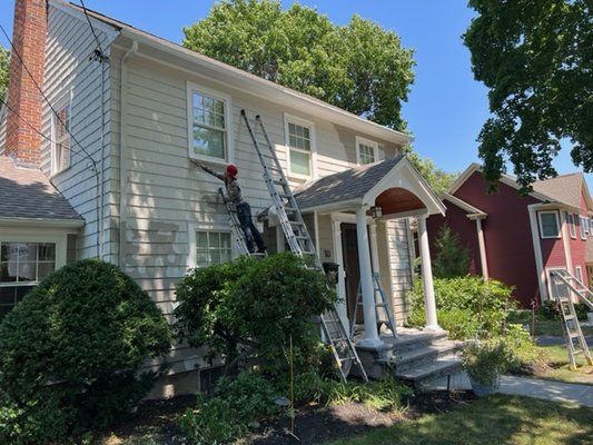 Person painting a two-story beige house using ladders on a sunny day. A red house is next door.