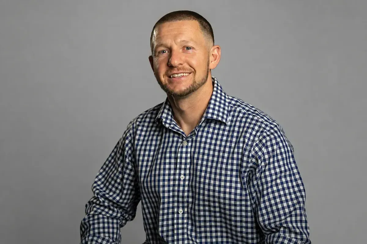 Smiling man in a blue checkered shirt seated against a gray background