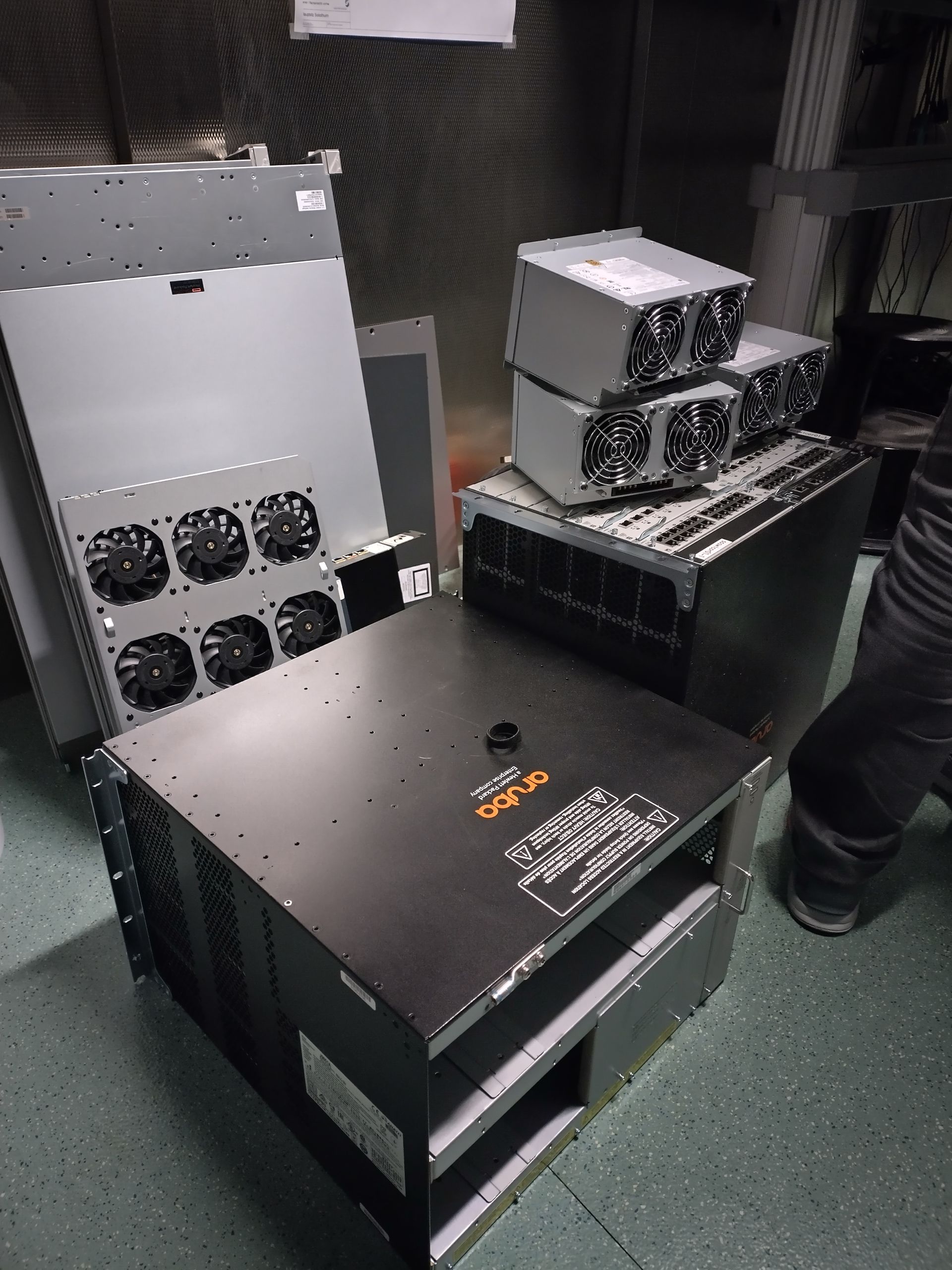 Gestapelte elektronische Geräte und schwarze Gehäuse in einem schwach beleuchteten Server- oder Technikraum.