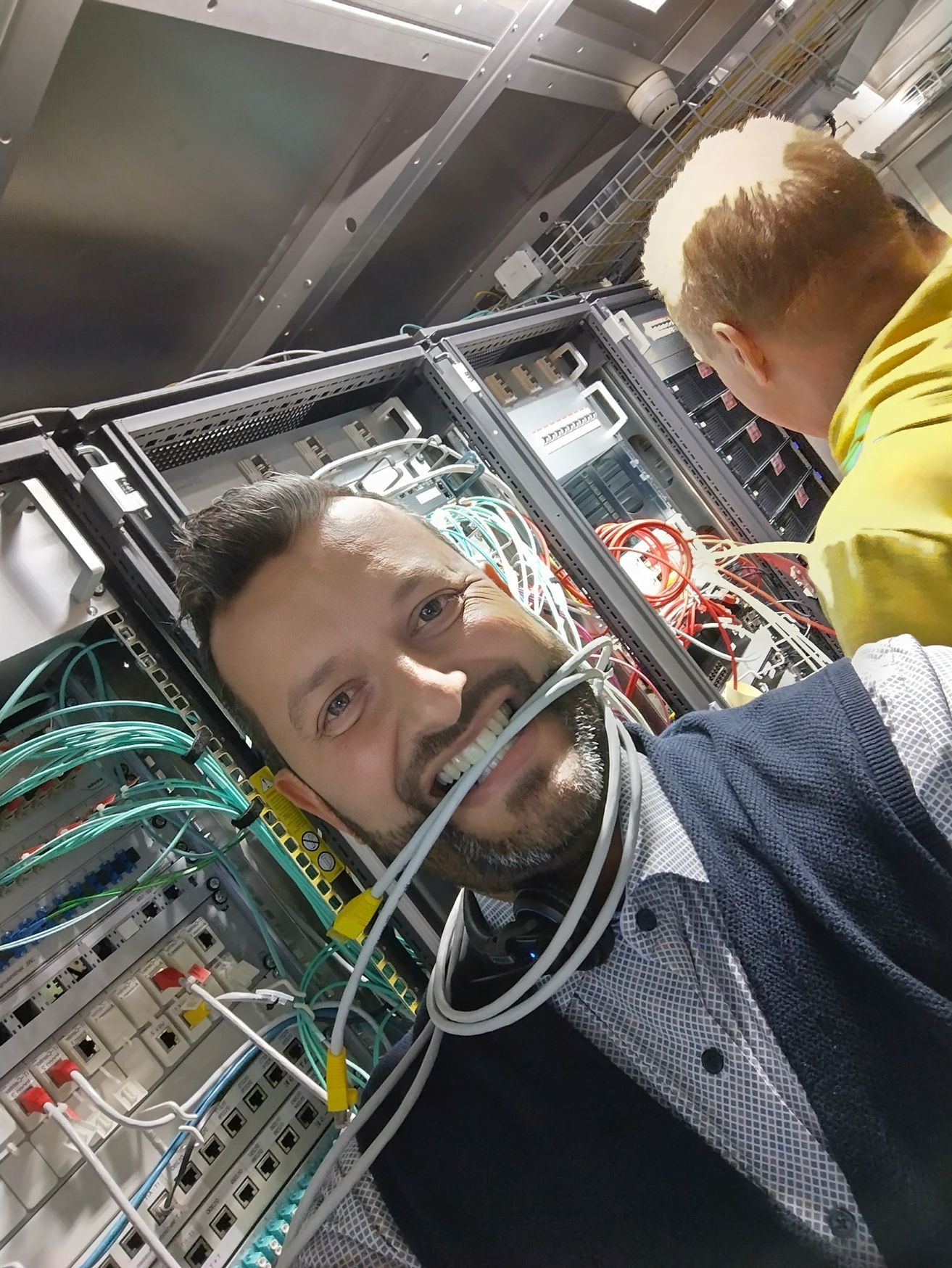 Ein Mann lächelt in einem Serverraum mit Kabeln; im Hintergrund arbeitet eine weitere Person.
