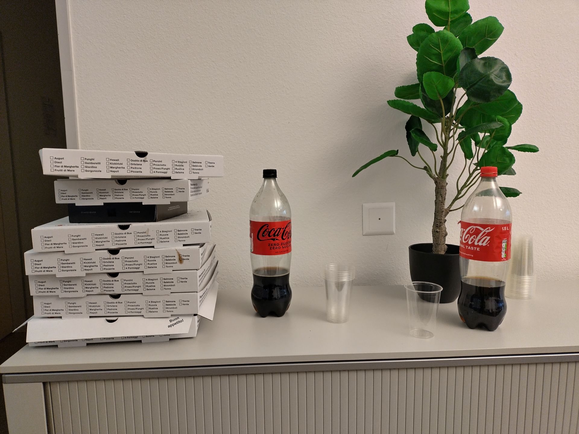 Stapel Pizzakartons mit zwei Coca-Cola-Flaschen und einer Topfpflanze auf einer Arbeitsplatte.