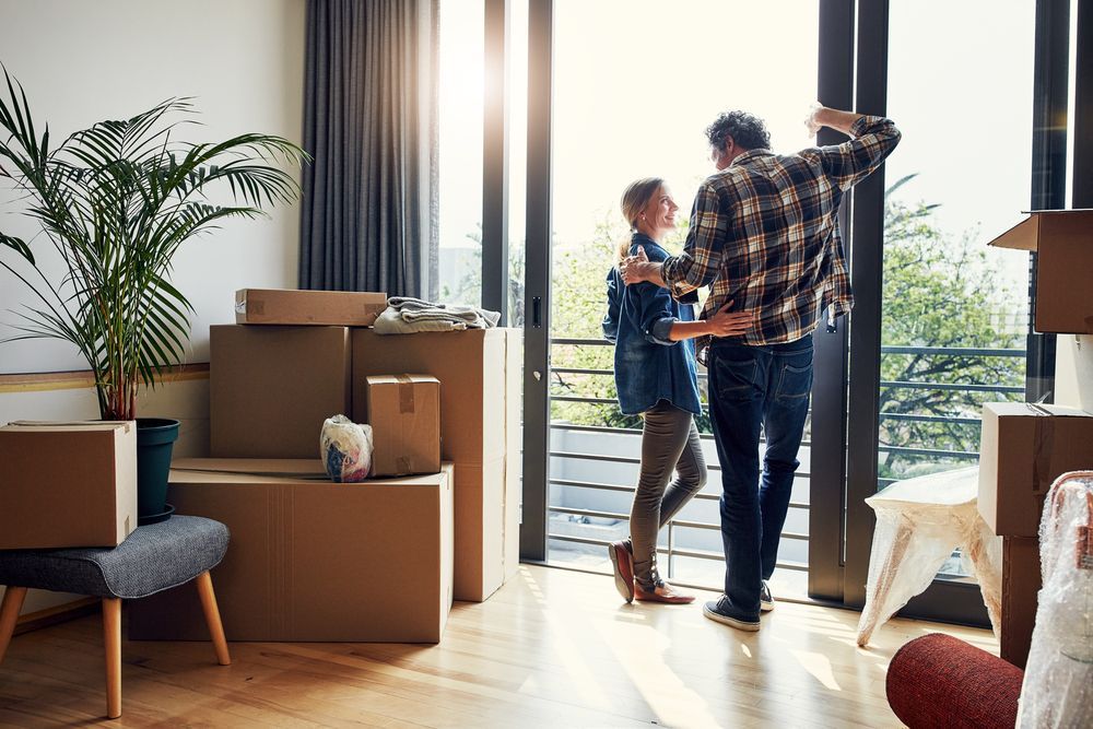 A man and a woman are dancing in a living room filled with cardboard boxes.