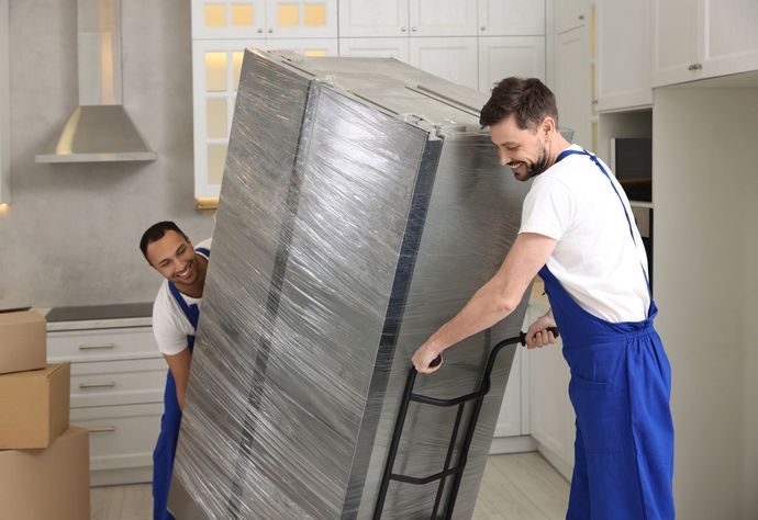 Two men are carrying a refrigerator on a cart in a kitchen.