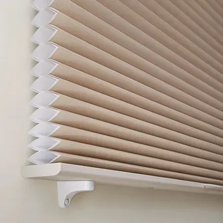 Tan pleated cellular shade partially covering a window, mounted on a white bracket.