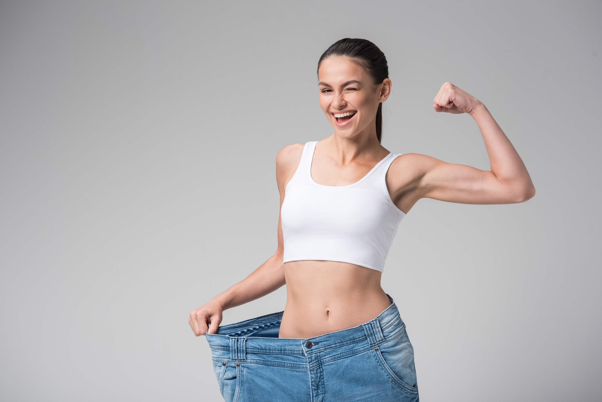 Smiling woman showing weight loss by holding oversized jeans and flexing her arm