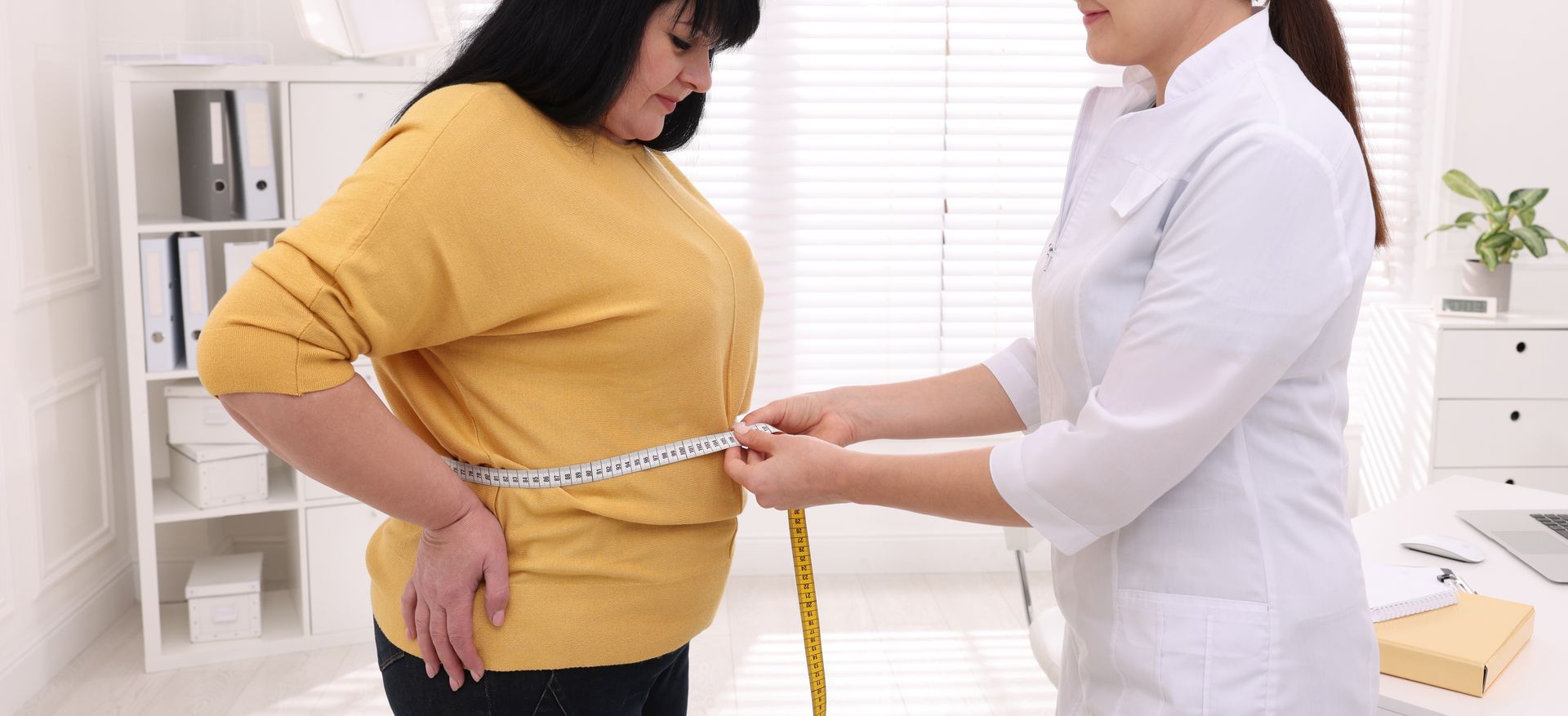 Nutritionist measuring overweight woman's waist with tape in clinic.
