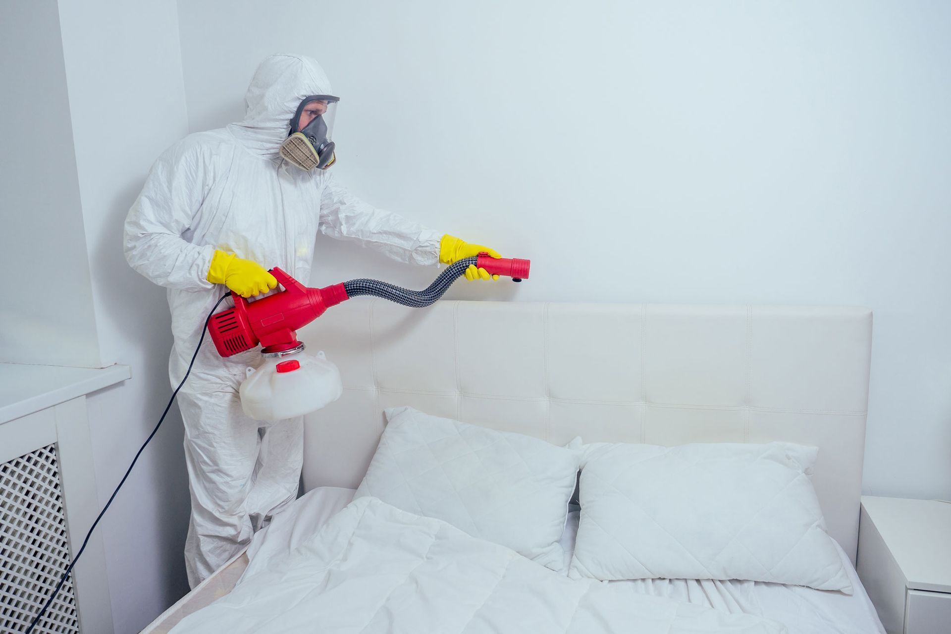 Worker in white protective suit spraying disinfectant over a bed in a white room.