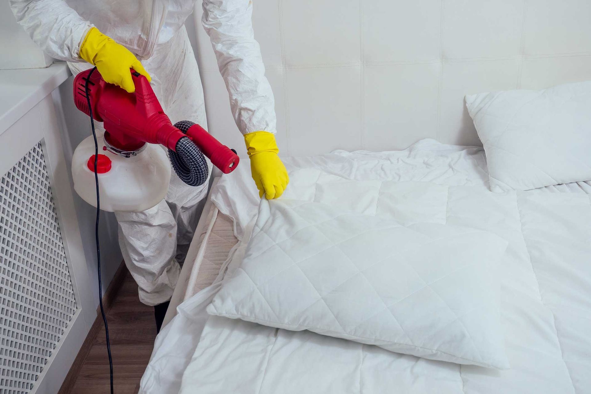 Person in white protective suit spraying disinfectant on a bed in a bedroom