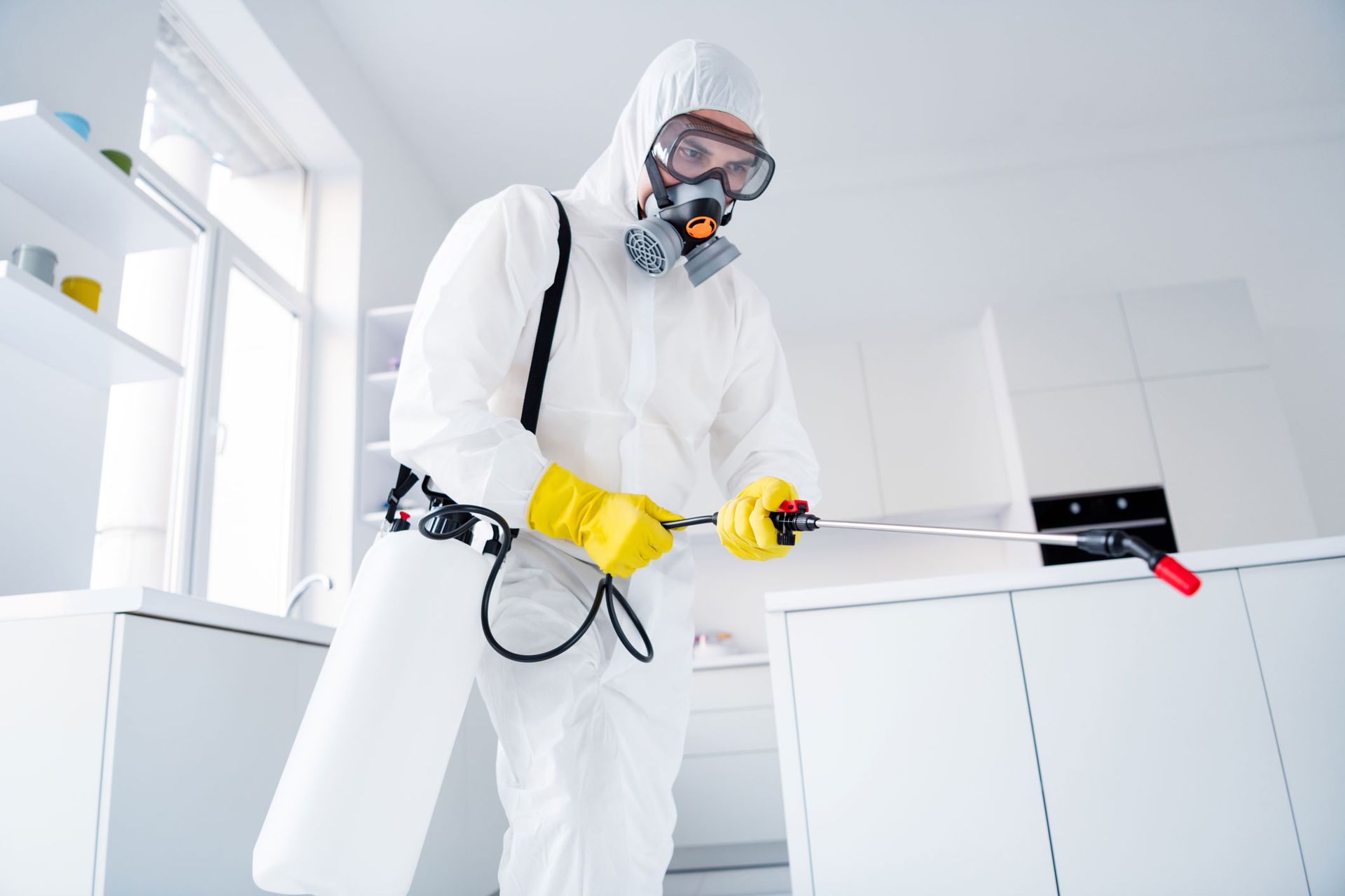 Worker in white protective suit and yellow gloves using a sprayer in a clean room near cabinets