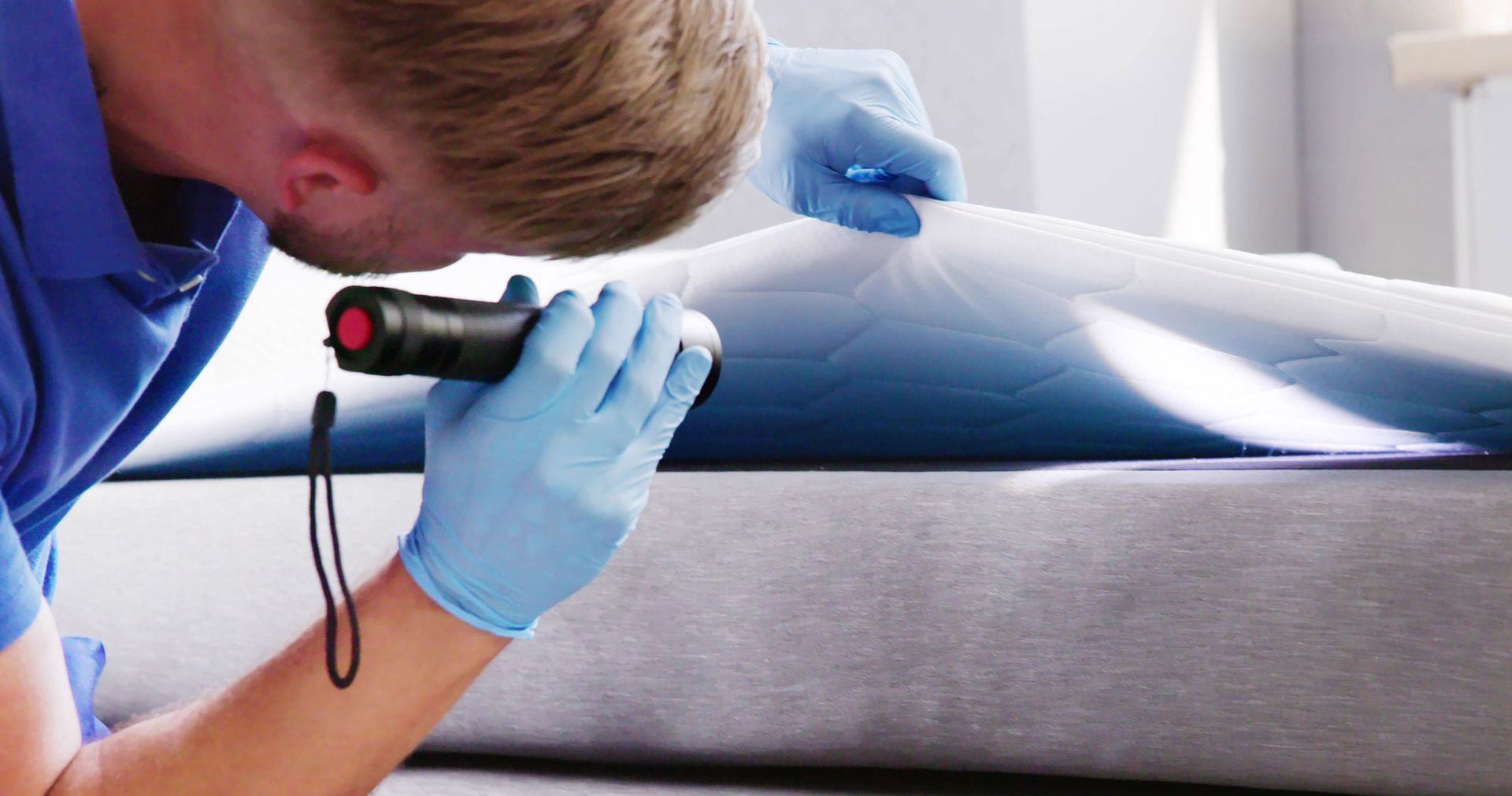 Technician using a flashlight to inspect a mattress seam with blue-gloved hands