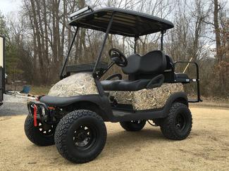 A golf cart with a camouflage paint job is parked in a dirt lot.