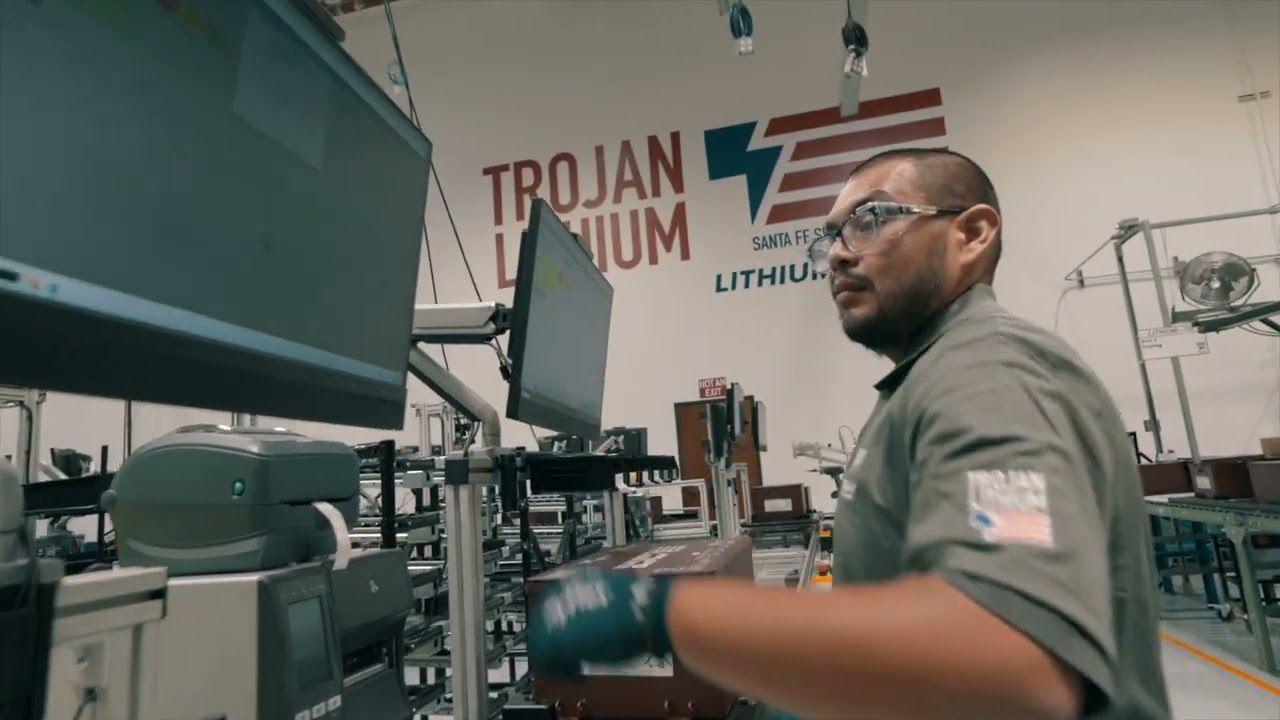 Man in a factory wearing safety glasses, checking a computer; Trojan Lithium logo visible.