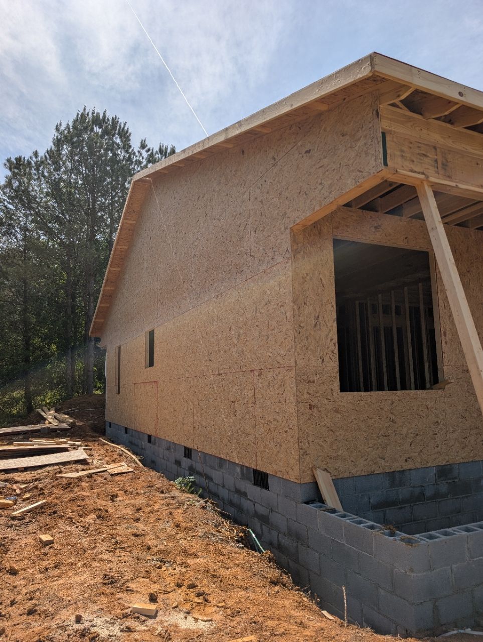 Partially built house with OSB walls, unfinished roof, and concrete block foundation on a dirt lot