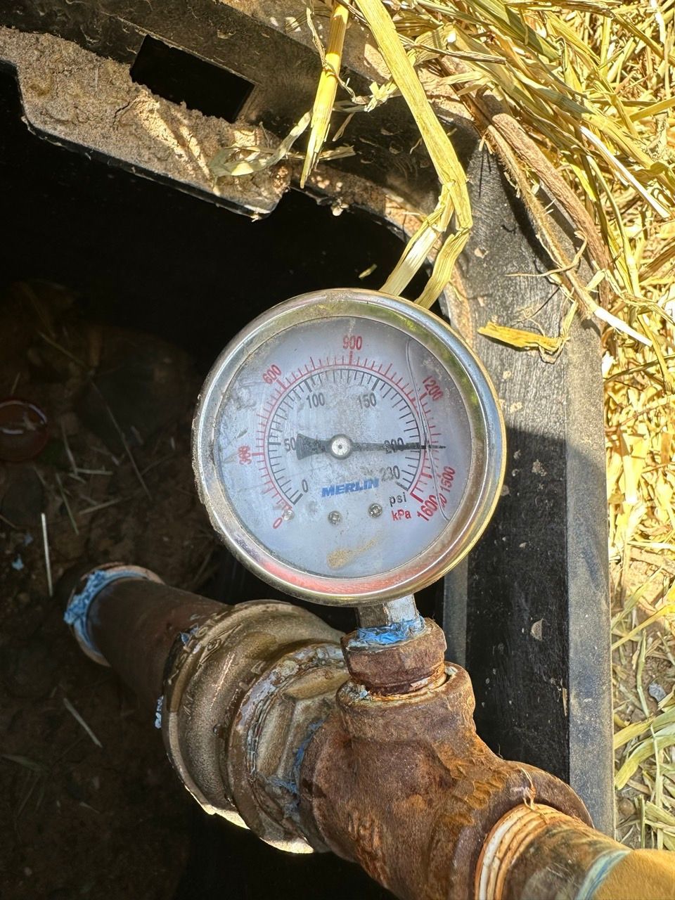 Close-up of a rusted pressure gauge on a pipe in a dark utility pit with yellow wires above.