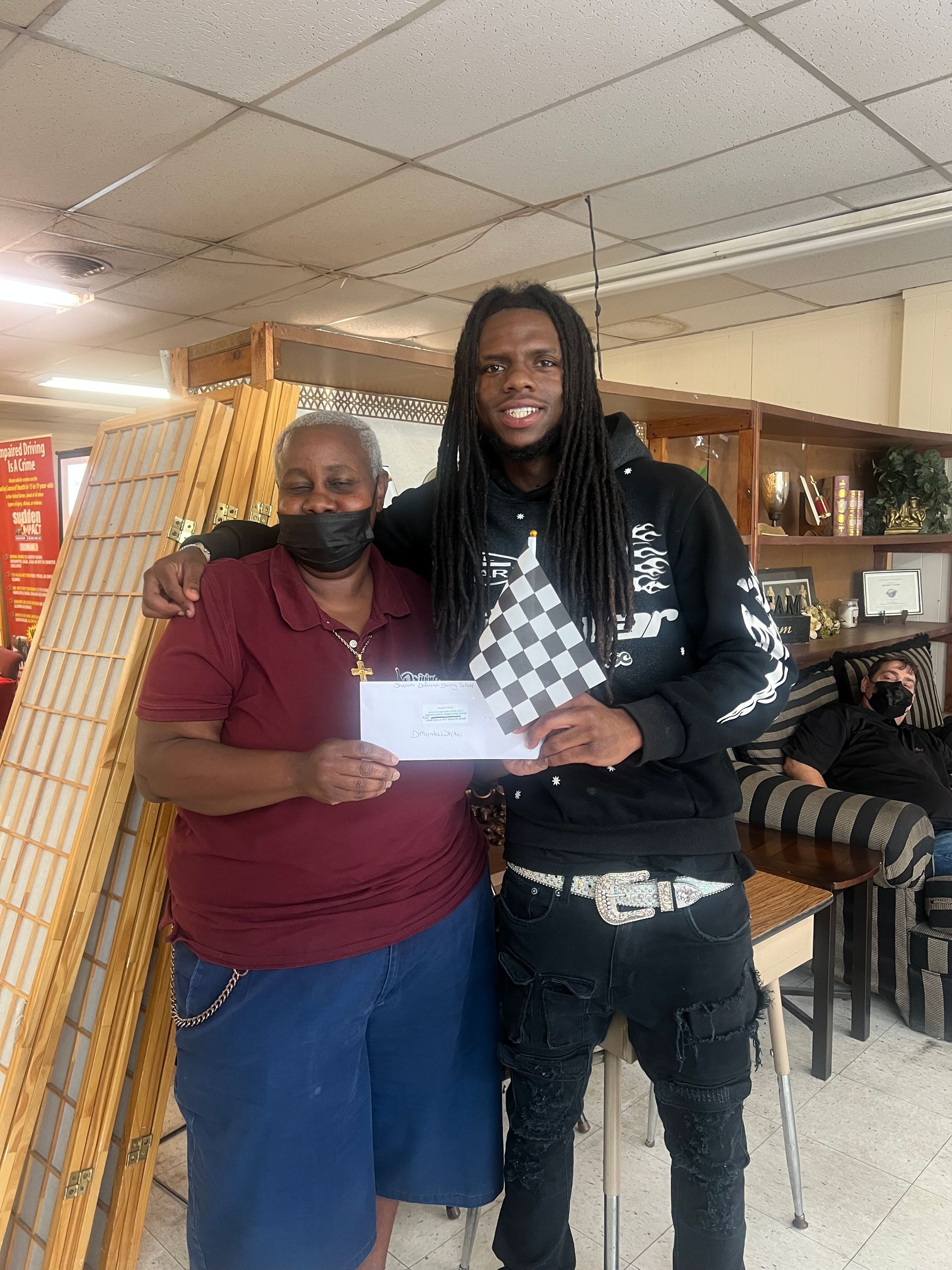 Woman and man hold a check. Man has long dreadlocks and a black jacket. Indoors with shelving.